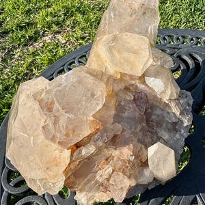 May include: A large, natural quartz crystal cluster with multiple points and a warm, amber color. The crystal formation has a rough, textured surface and sits on a decorative black metal table. The background shows green grass.