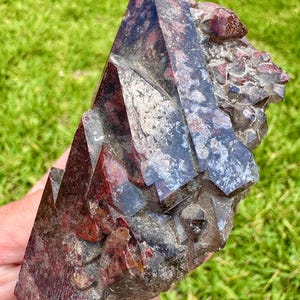 May include: A natural, raw mineral specimen featuring a cluster of dark red and gray crystals. The crystals have sharp, geometric shapes and a rough, textured surface. The mineral is held against a backdrop of green grass.