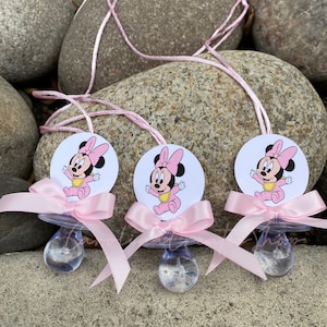 May include: Three baby pacifiers with a Minnie Mouse design. Each pacifier has a clear nipple, a pink bow, and a round tag with a cartoon image of Minnie Mouse. The pacifiers are connected by a pink string.