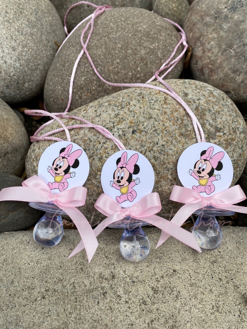 May include: Three baby pacifiers with a Minnie Mouse design. Each pacifier has a clear nipple, a pink bow, and a round tag with a cartoon image of Minnie Mouse. The pacifiers are connected by a pink string.