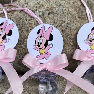 May include: Baby pacifiers featuring a Minnie Mouse design. Each pacifier has a clear nipple, a pink bow, and a circular image of Minnie Mouse in a pink outfit. A pink string is attached for hanging.