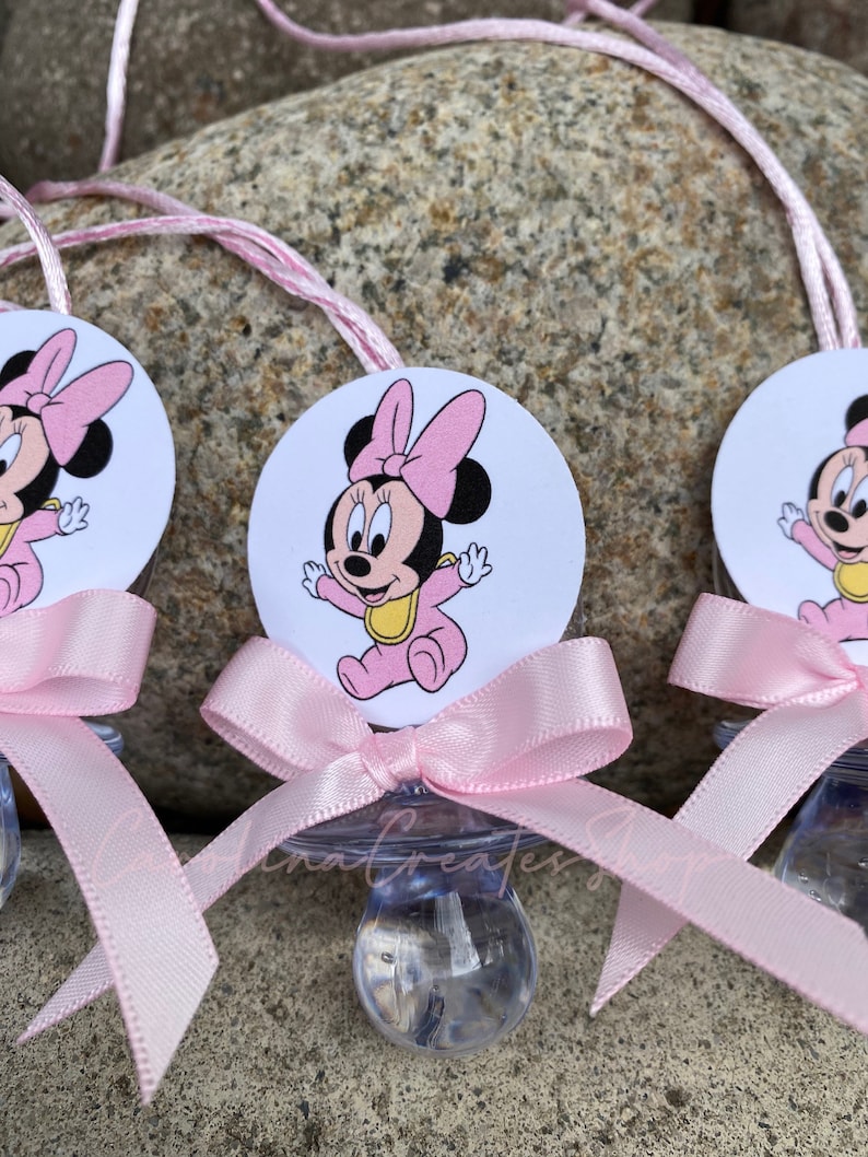 May include: Baby pacifiers featuring a Minnie Mouse design. Each pacifier has a clear nipple, a pink bow, and a circular image of Minnie Mouse in a pink outfit. A pink string is attached for hanging.