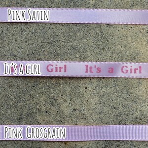 May include: Three ribbons in shades of pink are displayed. The top ribbon reads "PINK SATIN". The middle ribbon has the text "IT'S A GIRL" printed on it. The bottom ribbon reads "PINK GROSGRAIN".