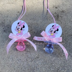 May include: Two baby pacifiers with pink ribbons and Minnie Mouse graphics. One pacifier is pink, the other is blue. The ribbons say "It's a girl" and "It's a boy". Each pacifier has a pink string attached.