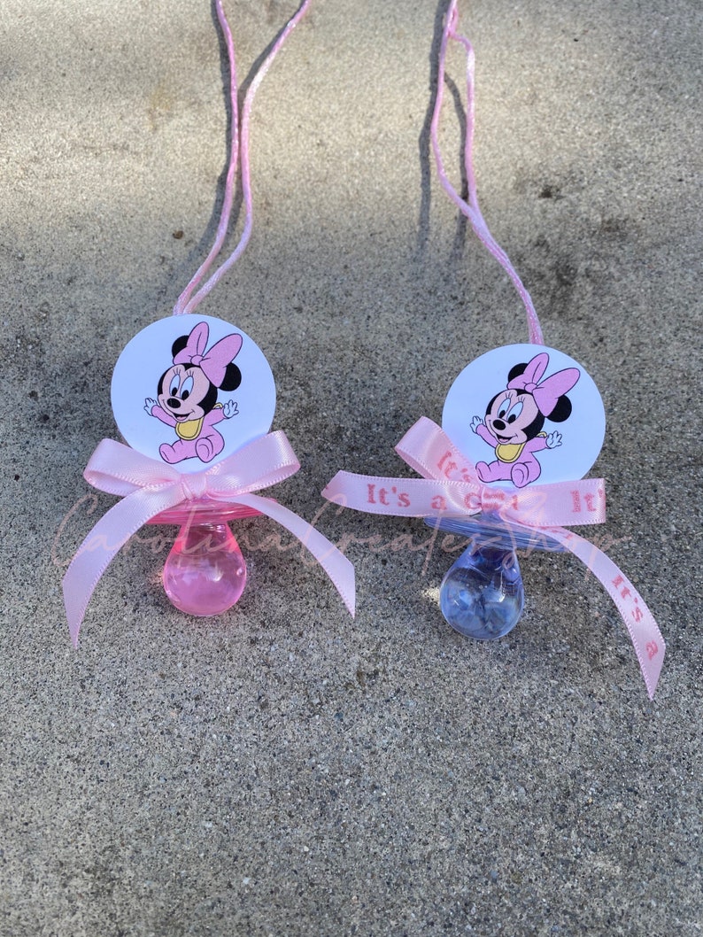 May include: Two baby pacifiers with pink ribbons and Minnie Mouse graphics. One pacifier is pink, the other is blue. The ribbons say "It's a girl" and "It's a boy". Each pacifier has a pink string attached.