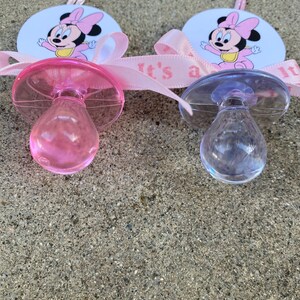 May include: Two baby pacifiers, one pink and one clear, with Minnie Mouse graphics. Each pacifier is attached to a pink ribbon that reads "It's a girl." The pacifiers are on a textured, gray surface.