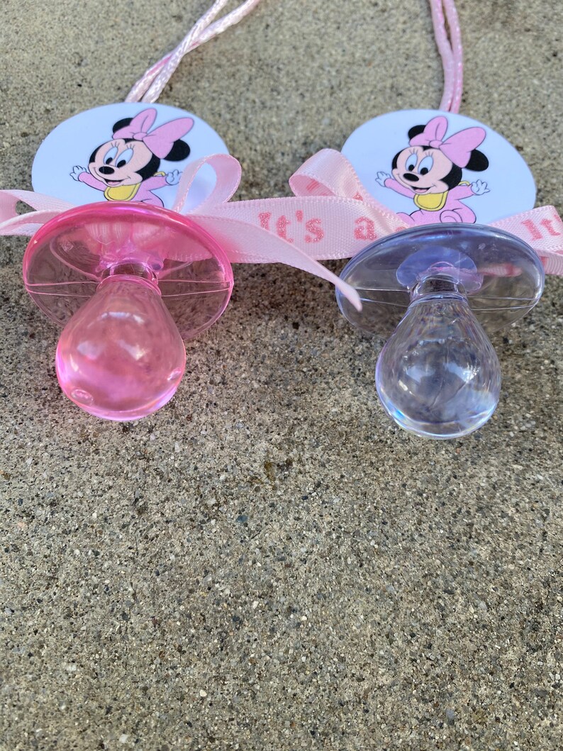 May include: Two baby pacifiers, one pink and one clear, with Minnie Mouse graphics. Each pacifier is attached to a pink ribbon that reads "It's a girl." The pacifiers are on a textured, gray surface.