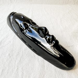 May include: A black ceramic sculpture of a face with a glossy finish. The sculpture is elongated and has a smooth, rounded shape.