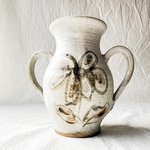 May include: A ceramic vase with a speckled off-white glaze and two side handles. The front features a hand-painted, abstract floral design in dark brown and muted earth tones. The base is unglazed, revealing the natural brown clay texture.