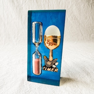 May include: A blue rectangular decorative piece featuring an hourglass with pink sand and a golden egg cup with the word "Eggs". Below the egg cup is a hen with the word "TIMER".