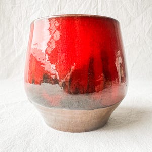 May include: A ceramic vase featuring a glossy, vibrant red glaze on the upper portion that transitions into a dark, smoky charcoal tone. The base is composed of an unglazed, matte brown clay material, creating a distinct contrast in texture.