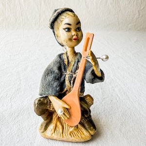 May include: A vintage ceramic figurine of a person playing a musical instrument. The figurine is painted in shades of brown, grey, and gold. The person is wearing a grey hat and a grey and gold robe. The instrument is a long, orange, wooden instrument with a round body.