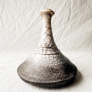 May include: A ceramic vessel with a wide, dark grey base that transitions into a narrow, tapered neck. The upper portion features a white glaze with a prominent black crackle pattern, while the lower section has a textured, dark grey finish.