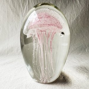 May include: A clear, oval paperweight featuring a pink jellyfish design. The jellyfish has a bell-shaped body and long, flowing tentacles. The paperweight is made of glass and rests on a white surface. The measurements are in centimetres.
