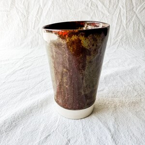 May include: A handcrafted ceramic vase with an organic shape. The vase has a dark, mottled glaze in brown, green, and red, with a white base. The interior rim has a glossy finish. This decorative piece is suitable for home decor. The height is approximately 20 cm.