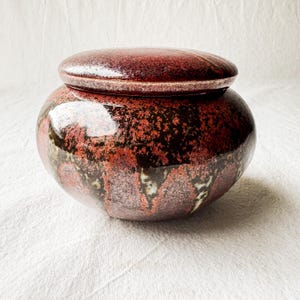May include: A round ceramic jar with a lid. The jar is glazed in shades of red, brown, and black, with a speckled pattern. The lid sits on top, creating a cohesive, decorative piece. The jar is likely used for storage.