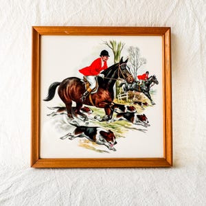 May include: A square framed print depicting a fox hunting scene with two riders in red coats on horseback and several hounds running across a grassy field. The artwork is enclosed in a simple light brown wooden frame with a white border.
