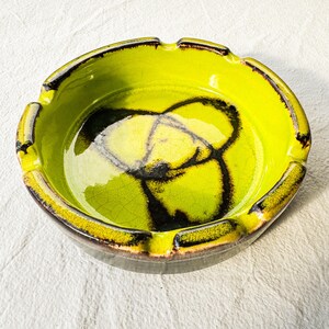 May include: A round ceramic ashtray featuring a vibrant lime green glaze with a dark brown crackle pattern. The circular dish has four evenly spaced notches along the rim, with dark brown accents highlighting the edges and the interior base.