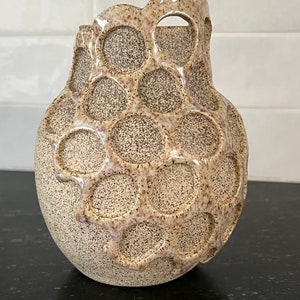 May include: A beige ceramic vase with a textured surface and a unique, circular, honeycomb-like design. The vase has a wide opening and a rounded base.