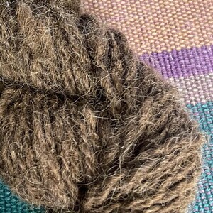 May include: A skein of brown wool yarn with a fuzzy texture. The yarn is wrapped and tied together with a green string.