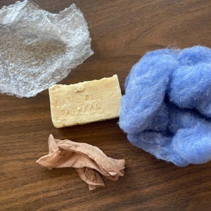 Puede incluir: Una pastilla de jabón de avena y lavanda, un trozo de plástico de burbujas, un manojo de lana azul y un trozo de tela marrón.