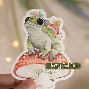 May include: A green frog sits on a red and white mushroom with white spots. The mushroom has a white stem and is on a white background. The frog has a small snail on its back and a butterfly on its head. The text "Ivory Owl Co" is on the mushroom.