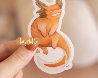 Peut inclure: Un autocollant représentant un axolotl de dessin animé de couleur orange et jaune, avec des branchies vertes, sur un fond blanc. L'autocollant porte l'inscription "Ivory Owl Co.".