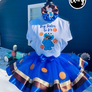 Peut inclure: Une tenue sur le thème de Cookie Monster, comprenant un t-shirt blanc avec l'inscription "Big Sister To K3" et un motif Cookie Monster. La tenue comprend une jupe tutu bleue avec des motifs de biscuits, un nœud assorti et un bandeau marron.