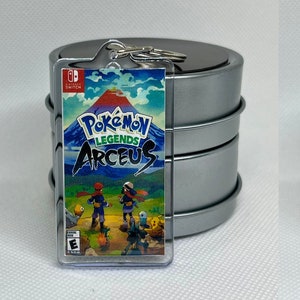 May include: A clear plastic keychain featuring the front and back of the Pokemon Legends Arceus video game case for the Nintendo Switch. The front of the case features a mountain range with a red volcano, two characters, and the game title. The back of the case features a variety of Pokemon characters and the game logo.
