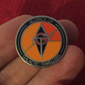 Puede incluir: Un pin de plata con un diseño negro y naranja. El pin presenta un triángulo estilizado con las letras "GT" en su interior. El texto "ALLIANCE OF GALACTIC TRAVELLERS" está escrito alrededor del exterior del pin.