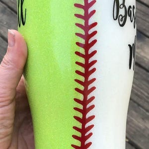 May include: A baseball-themed tumbler with a glittery lime green and white design. The tumbler features a red stitch pattern down the center, with the word "Baseball" and a name in black script. The tumbler is held by a hand.