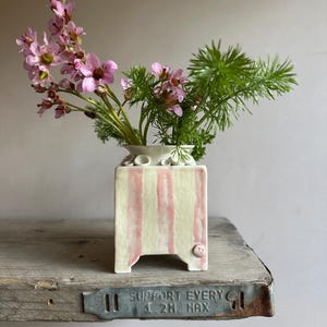 May include: A small, square ceramic vase with pink vertical stripes and a small button detail on the side. The vase holds a bouquet of pink flowers and green foliage. The vase is sitting on a rustic wooden shelf.