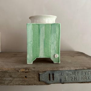 May include: A ceramic decorative piece with a white top and a green, vertically striped body. The square-shaped base has small feet and a button detail. The piece is displayed on a wooden shelf.