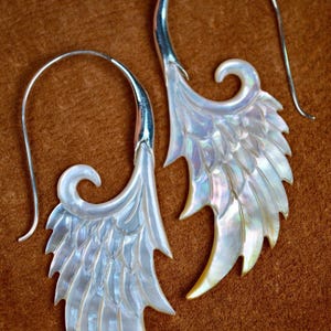 May include: A pair of iridescent, wing-shaped earrings. The earrings feature a silver hook and a detailed, layered design with a pearlescent sheen. The wings have a curved, feather-like pattern, with a pointed edge.