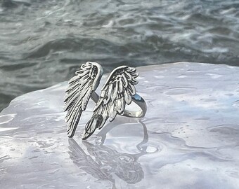 May include: A silver-colored ring featuring a detailed wing design. The ring's wings are spread open, showcasing intricate feather detailing. The ring is set against a reflective, wet surface with a blurred water background.