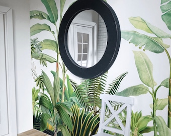 May include: A black framed round mirror with a tropical leaf wallpaper backdrop. The wallpaper features large green leaves and a white background. A white chair is in the foreground.