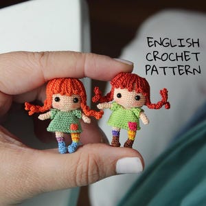 Pode incluir: Duas pequenas bonecas de crochê feitas à mão, com cabelo ruivo e vestidos verdes. As bonecas têm meias arco-íris e são seguradas em uma mão. O texto "ENGLISH CROCHET PATTERN" está ao fundo.