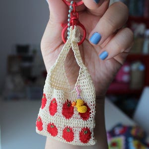 Könnte beinhalten: Ein cremefarbenes, gehäkeltes Miniaturtäschchen mit einem wiederkehrenden Muster aus roten Erdbeeren mit grünen Stielen. Das Täschchen ist an einem roten Kunststoffschlüsselring befestigt und hat einen kleinen, gelben Häkel-Entenanhänger.