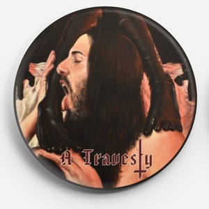Può includere: Un bottone bianco e nero con un dipinto di un uomo con i capelli lunghi e la barba. L'uomo è tenuto da due mani. Il testo "A Travesty" è stampato in rosso sul bottone.