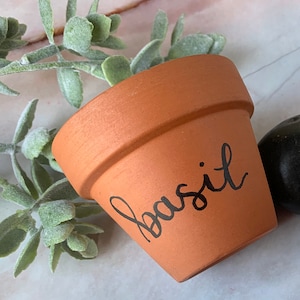 Puede incluir: Una maceta de terracota con la palabra "albahaca" escrita en caligrafía negra en el lateral.