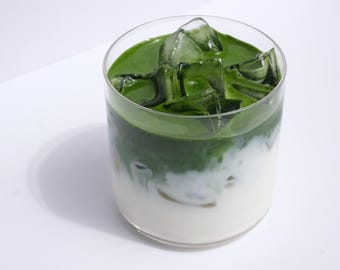 Può includere: Un matcha latte rinfrescante in un bicchiere trasparente. La bevanda è stratificata con una base bianca cremosa, un vivace strato verde matcha e cubetti di ghiaccio. Il bicchiere è su una superficie bianca, con un'ombra laterale. Misure in cm e ml.