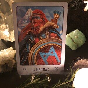 Peut inclure: Une carte avec un bouclier bleu et rouge avec un symbole de rune. La carte est d'un jeu de runes et est &eacute;tiquet&eacute;e "20. Mannaz".