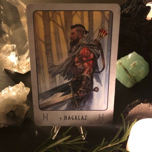 Peut inclure: Une carte de tarot avec un fond gris et une bordure blanche. La carte repr&eacute;sente un homme barbu avec une &eacute;p&eacute;e dans un d&eacute;cor de for&ecirc;t. La carte est intitul&eacute;e "Hagalaz" avec un symbole de rune.