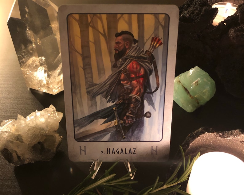 Peut inclure: Une carte de tarot avec un fond gris et une bordure blanche. La carte repr&eacute;sente un homme barbu avec une &eacute;p&eacute;e dans un d&eacute;cor de for&ecirc;t. La carte est intitul&eacute;e "Hagalaz" avec un symbole de rune.