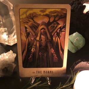 Peut inclure: Une carte de tarot avec un fond dor&eacute; et une image de trois femmes portant des robes &agrave; capuchon. La carte est intitul&eacute;e "39. Les Nornes".