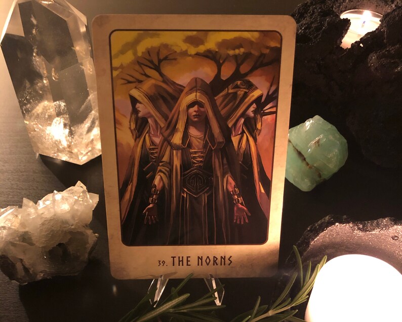 Peut inclure: Une carte de tarot avec un fond dor&eacute; et une image de trois femmes portant des robes &agrave; capuchon. La carte est intitul&eacute;e "39. Les Nornes".