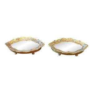May include: Two gold-colored decorative trays with ornate floral designs and mirrored surfaces. Each tray has three small feet.