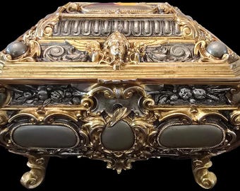 May include: An ornate, gold and silver, decorative box with intricate carvings and a raised, central design. The box has four feet and a hinged lid.