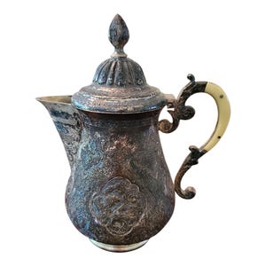 Puede incluir: Una tetera de plata antigua con un diseño ornamentado y un asa de hueso. La tetera tiene un cuerpo bulboso y una tapa abovedada.