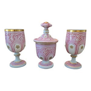 May include: A set of three antique glass pieces with pink and white enamel decoration. The set includes two goblets and a lidded jar.